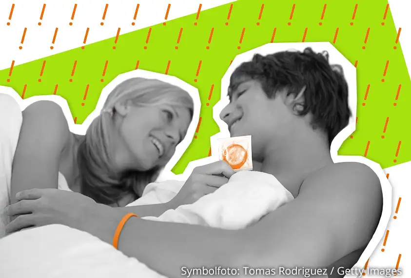 L&auml;chelndes Paar im Bett, die Frau h&auml;lt ein Kondom in der Hand.